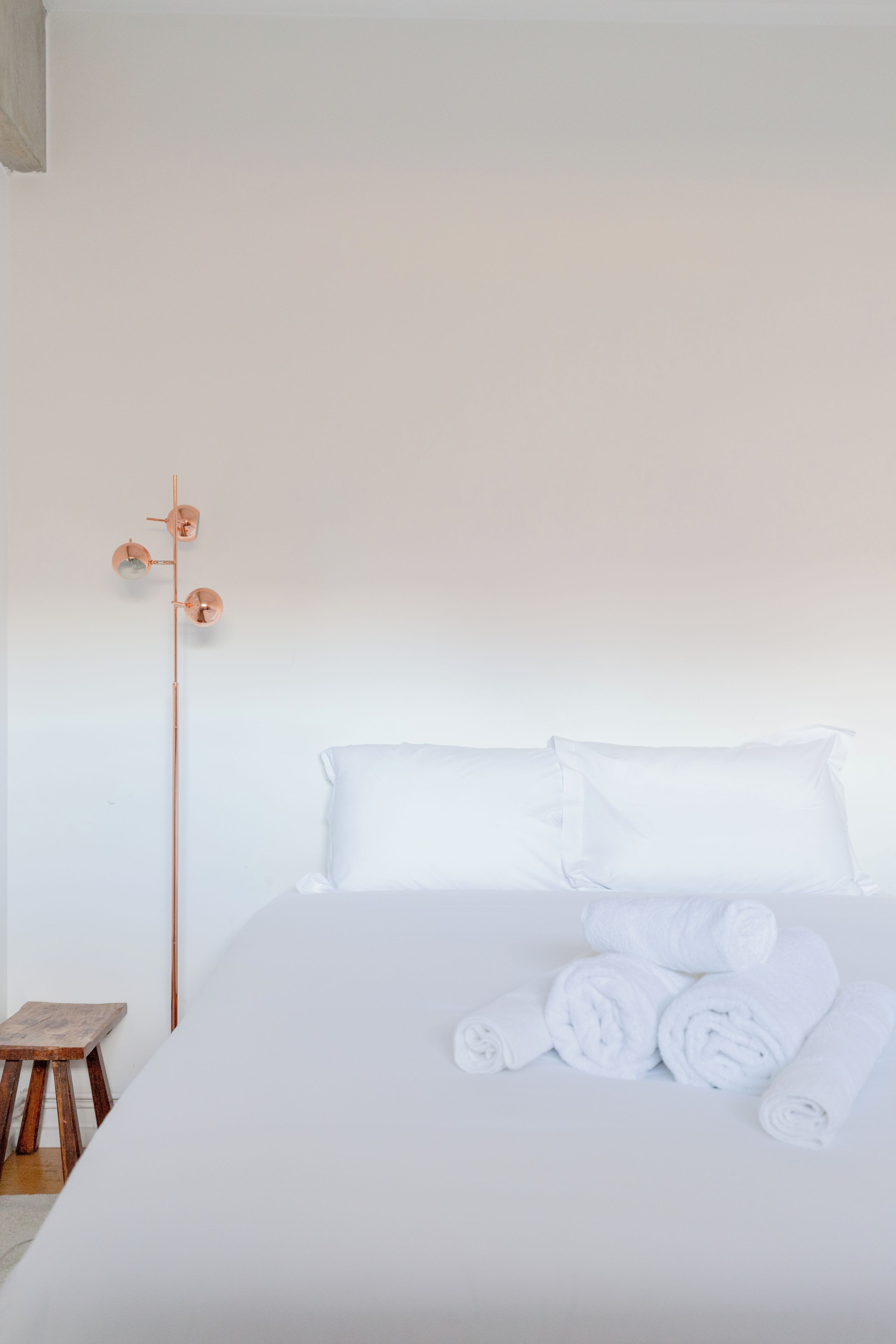A neatly made bed with white linens and rolled towels, a wooden stool, and a tall floor lamp with a single light fixture.