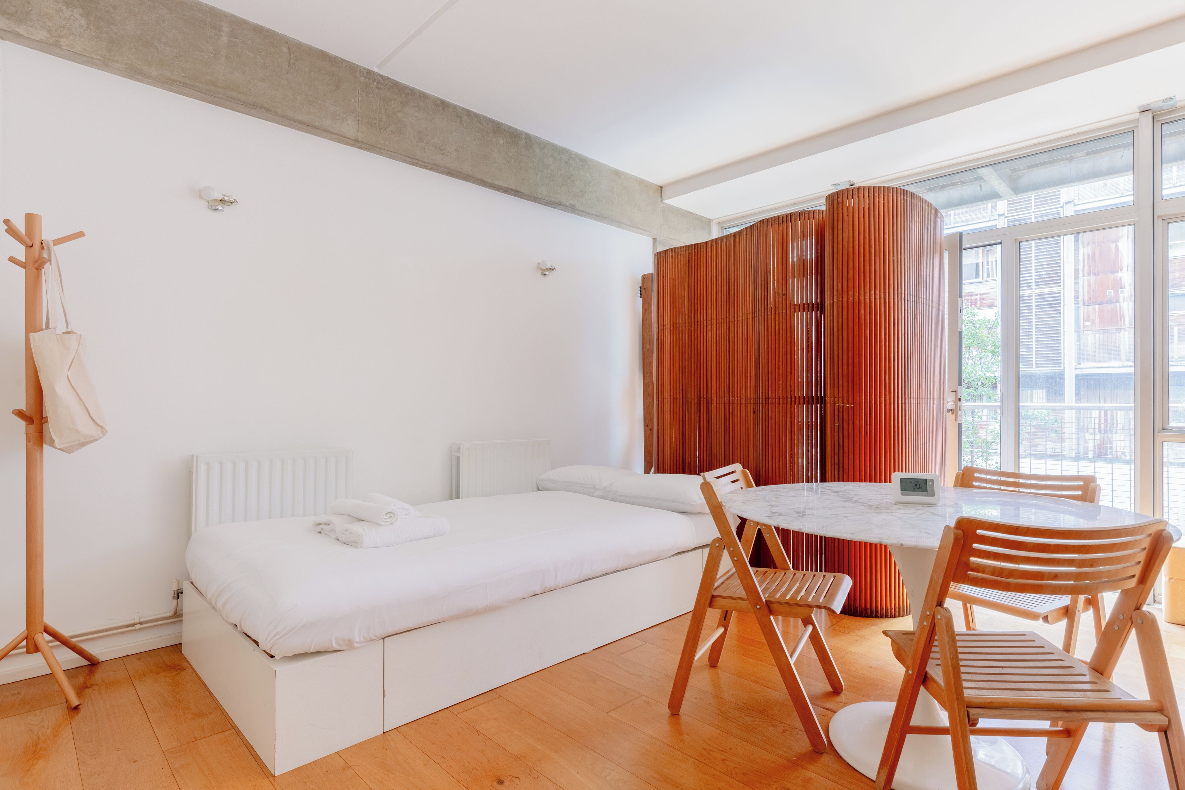 A modern room with a bed, a dining table with chairs, a wooden coat rack, and a window with blinds.