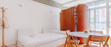 A modern room with a bed, a dining table with chairs, a wooden coat rack, and a window with blinds.