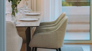 A dining area with a table set for two, a vase with flowers, and a view of the outdoors through a large window. - Luxurious Stylish 2 Bd Villa in Voula (Vari-Voula-Vouliagmeni)