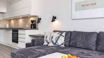Een moderne woonkamer met een grijze bank, een bijzettafeltje met boeken en fruit, en een keuken op de achtergrond.