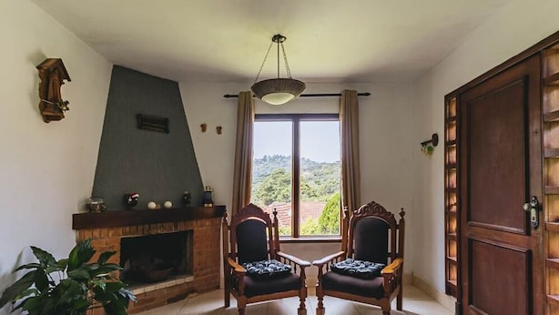 Un salon avec une cheminée, deux fauteuils, une table et une vue sur la verdure à l’extérieur.