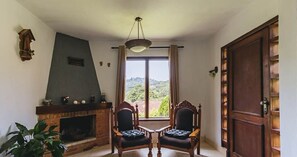 Un salón con chimenea, dos sillones, una mesa y vistas al exterior con vegetación.