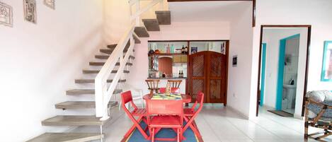 Un coin repas avec une table et des chaises rouges, un escalier et une cuisine en arrière-plan.