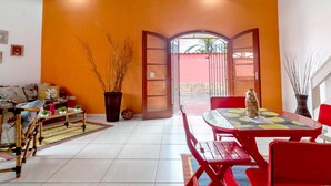 A living room with a sofa, a dining table with red chairs, a potted plant, and a view of the outside.