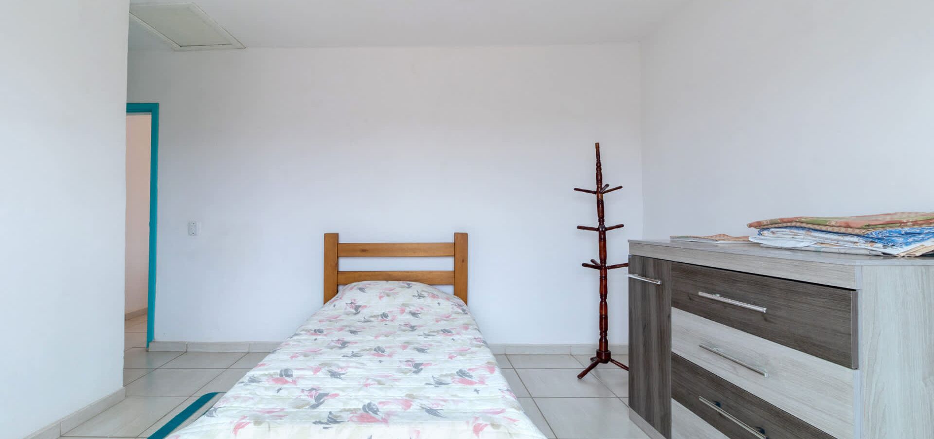 Una habitación pequeña y limpia con una cama individual, cabecera de madera, perchero y cómoda con una manta doblada encima.