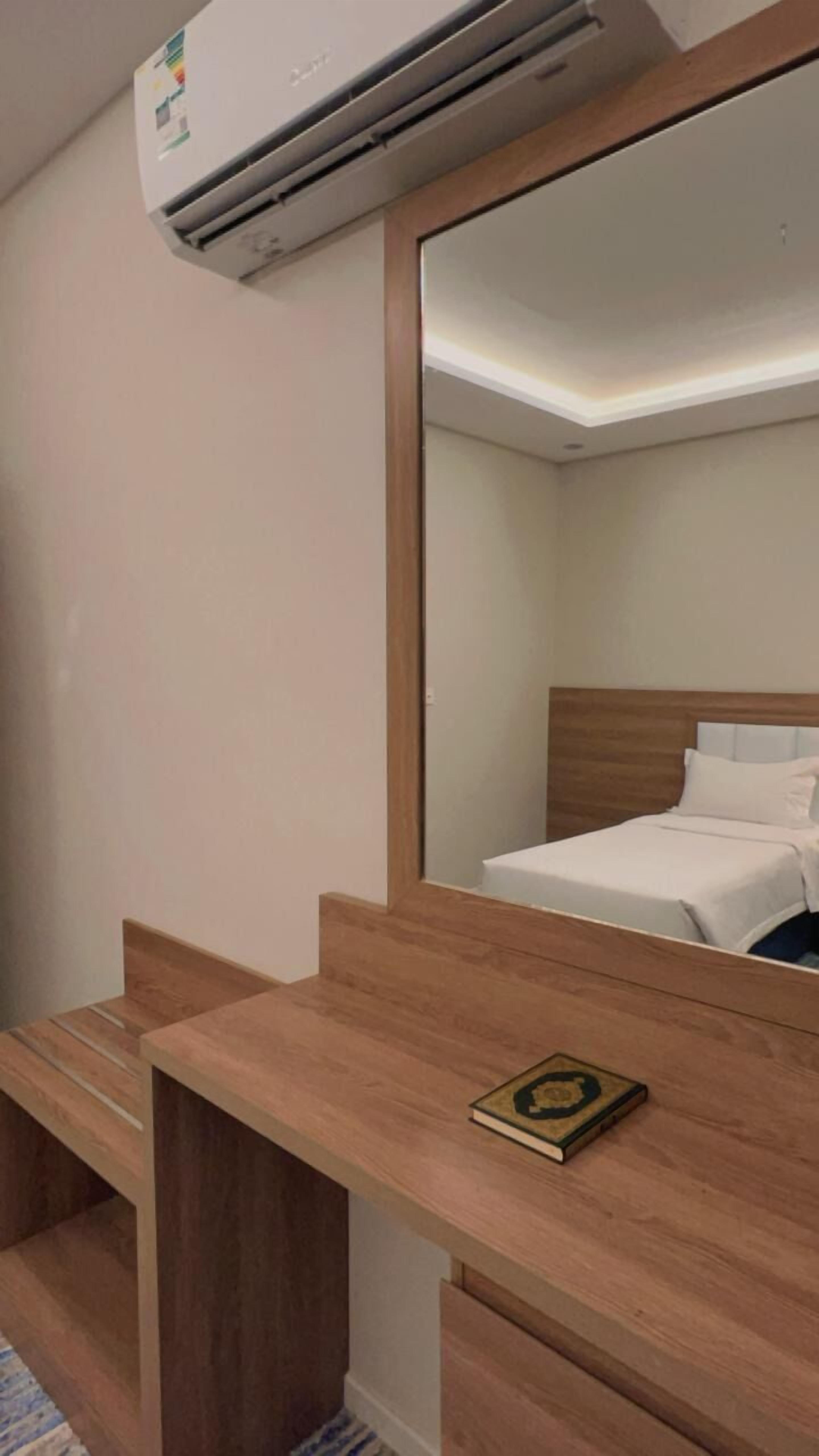 A hotel room with a wooden desk, a book, a bed with white bedding, and a ceiling-mounted air conditioner.