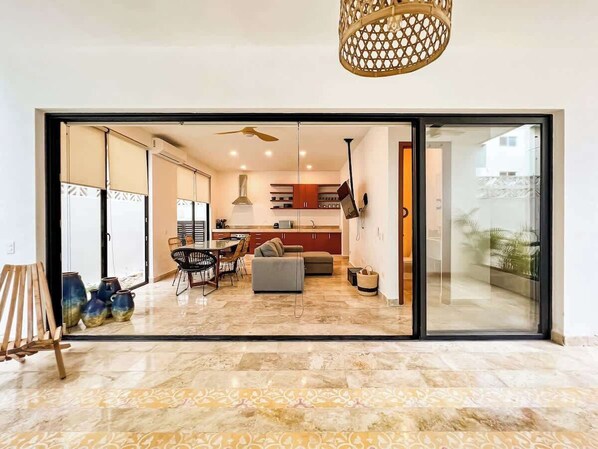 A spacious living area with a sliding glass door leading to a balcony, a dining area with chairs and a table, and a kitchen in the background.