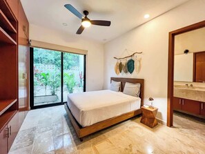 A bedroom with a bed, a ceiling fan, a nightstand, a bathroom with a sink, and a view of greenery outside.