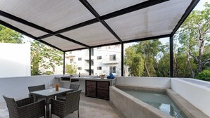 Un espacio al aire libre techado con una mesa y sillas, una piscina pequeña y vistas de edificios y árboles circundantes.