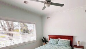 Une chambre avec un lit en bois, un ventilateur de plafond, une fenêtre avec des stores et une table de chevet sur laquelle se trouve un vase.