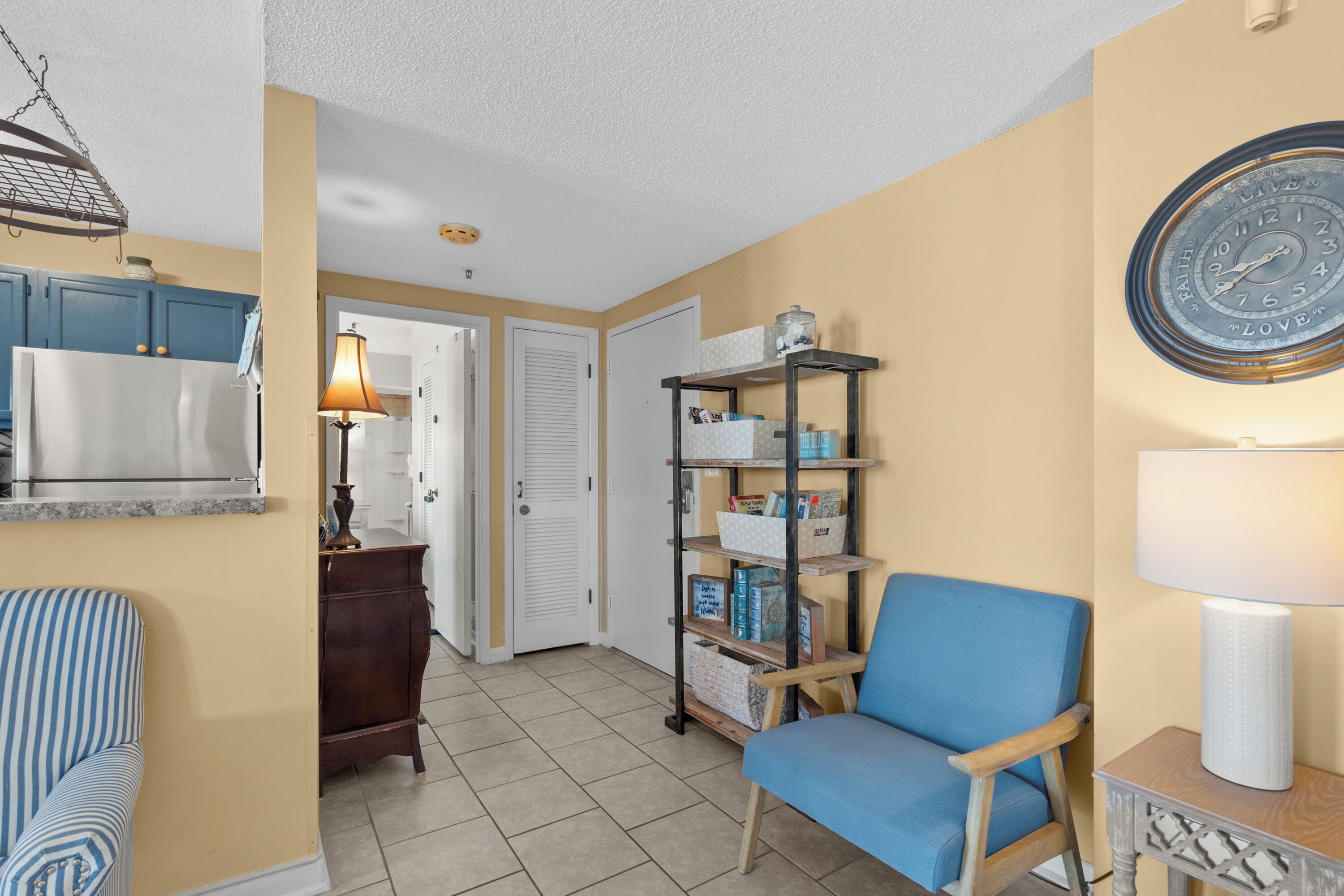 A living room with a blue armchair, a bookshelf, and a clock on the wall.