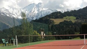 Een tennisbaan met een net, omringd door een groene hekken, en een bergachtig landschap met besneeuwde toppen op de achtergrond.
