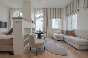 A modern living room with a sofa, a round table, and a chair. There is a television mounted on the wall, and a window with curtains. - Apartments Hotel Stadt Hamburg (Sylt)