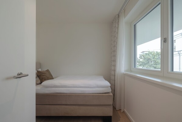 Un petit lit, bien fait, dans une chambre d’angle avec une fenêtre, une porte et un mur blanc.
