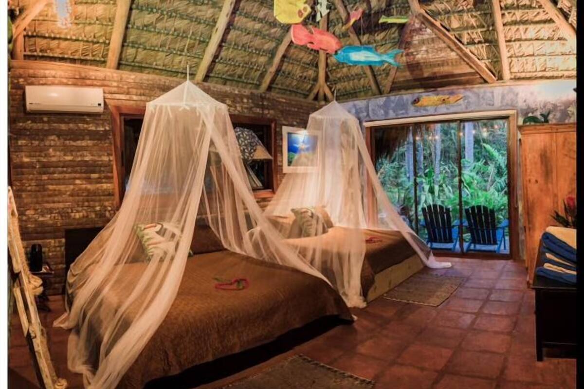 A rustic room with a thatched roof, two beds with mosquito nets, a fireplace, and a view of greenery through a glass door.