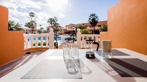 Een balkon met een tafel gedekt voor twee personen, met uitzicht op een zwembadgedeelte met palmbomen en resortgebouwen op de achtergrond.
