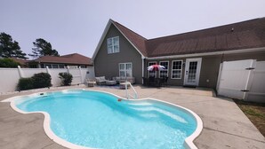 A swimming pool with a white edge, a house with a brown roof, and a white fence.