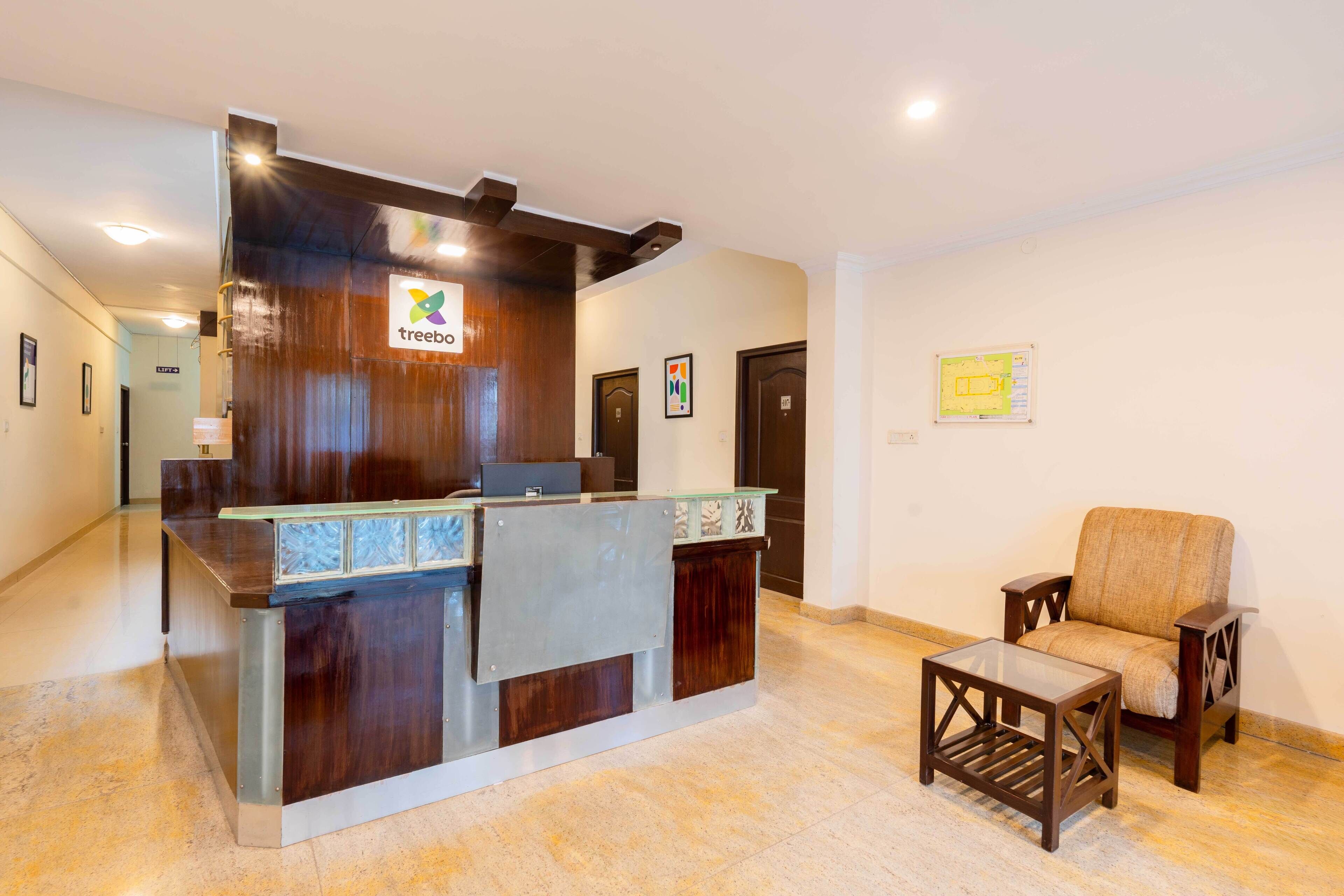 Bancada de recepção com logotipo, área de espera com uma cadeira e uma pequena mesa, e corredor com portas e quadros.