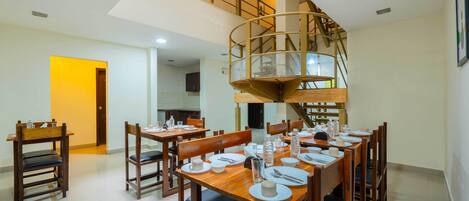 Un espace repas avec des tables et des chaises en bois, un escalier en colimaçon et une cuisine en arrière-plan.