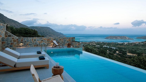 An infinity pool with lounge chairs, a stone wall, and a view of the sea and hills.