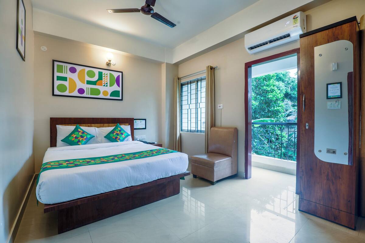 A hotel room with a bed, a chair, a window with a view of greenery, and an air conditioning unit.