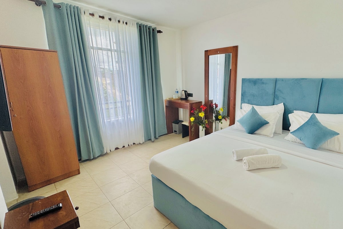A hotel room with a bed, a wooden wardrobe, a desk with a chair, a mirror, and a window with curtains.