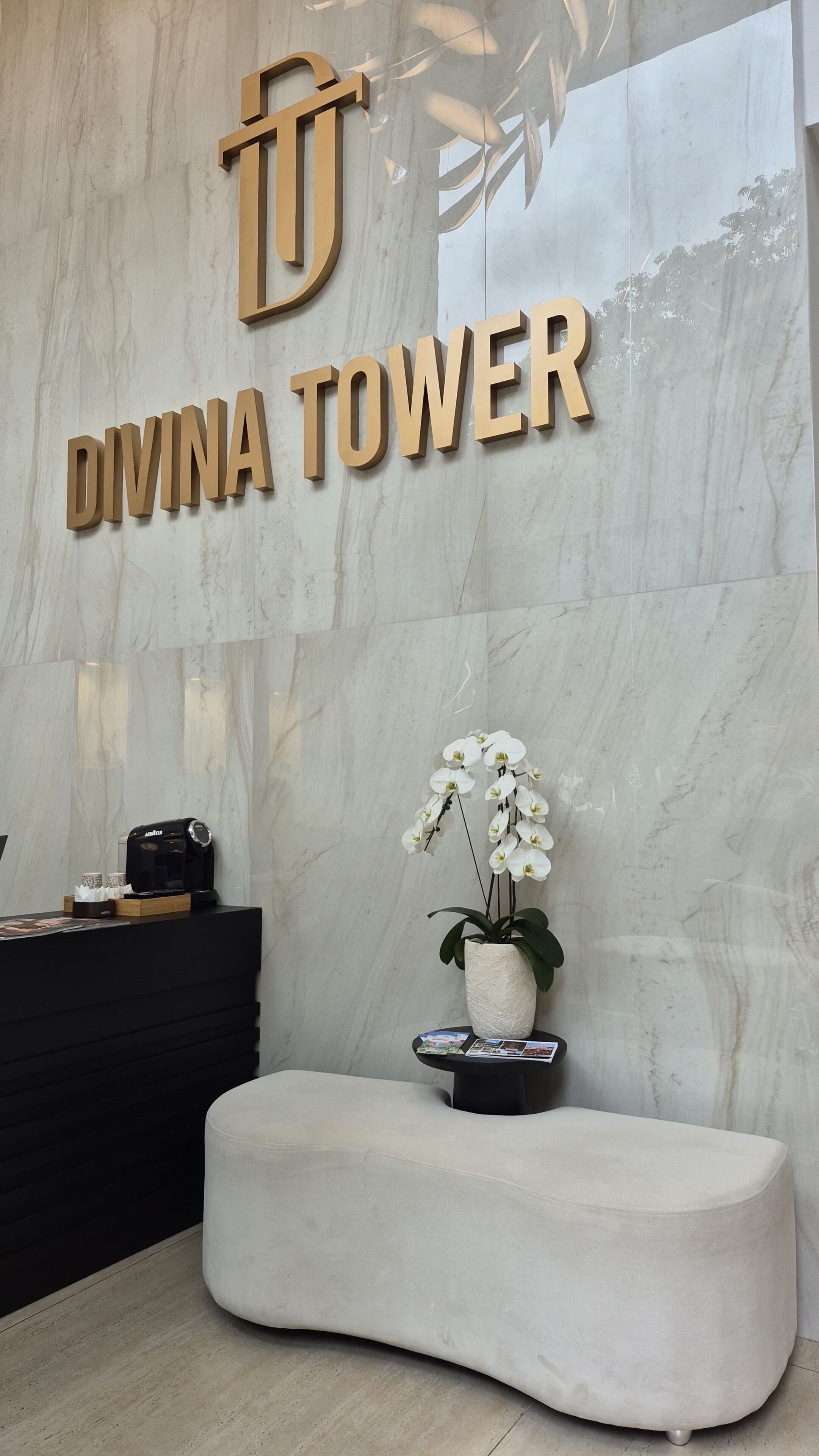 Una zona di accoglienza con parete in marmo, un cartello che riporta 'DIVINA TOWER', una scrivania reception nera, una panca bianca e un'orchidea in vaso.