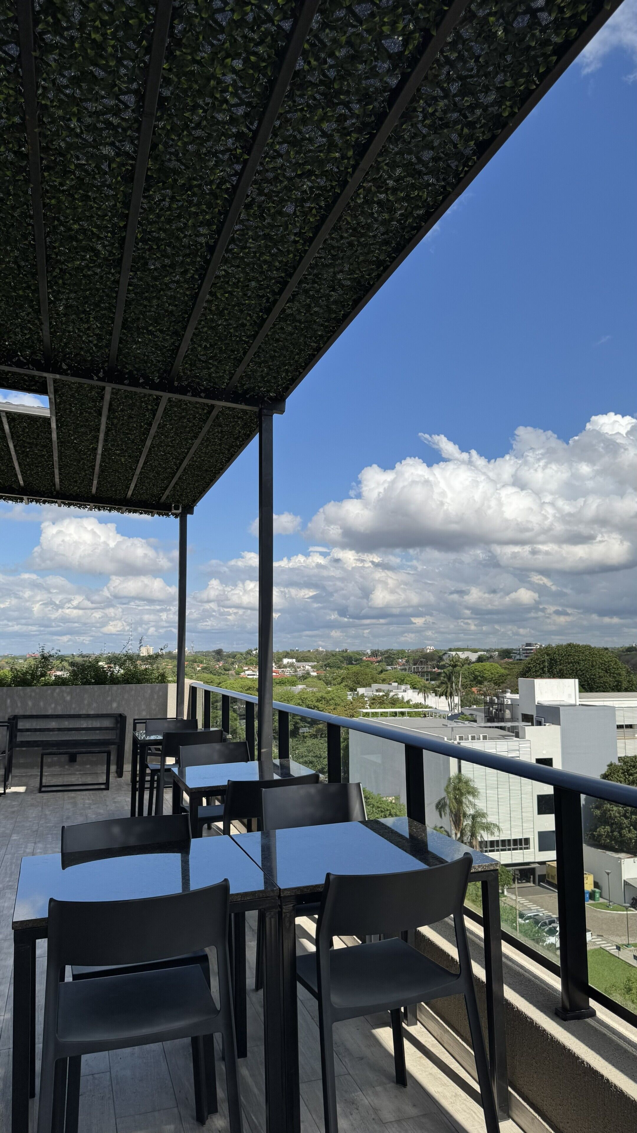 A covered outdoor seating area with tables and chairs, overlooking a cityscape with buildings and clouds.