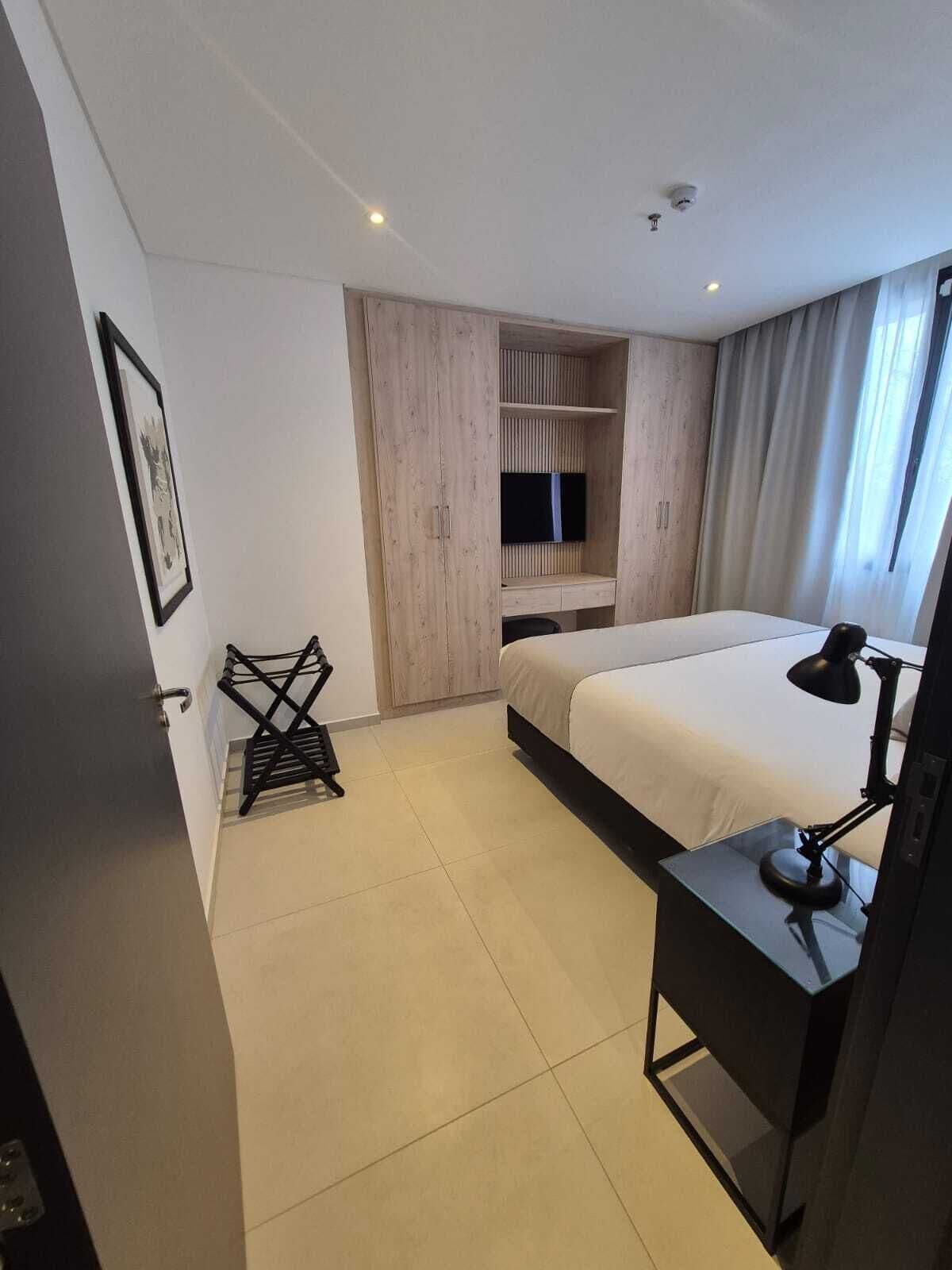 A hotel room with a bed, a desk with a lamp, a TV mounted on a wooden cabinet, and a framed picture on the wall.