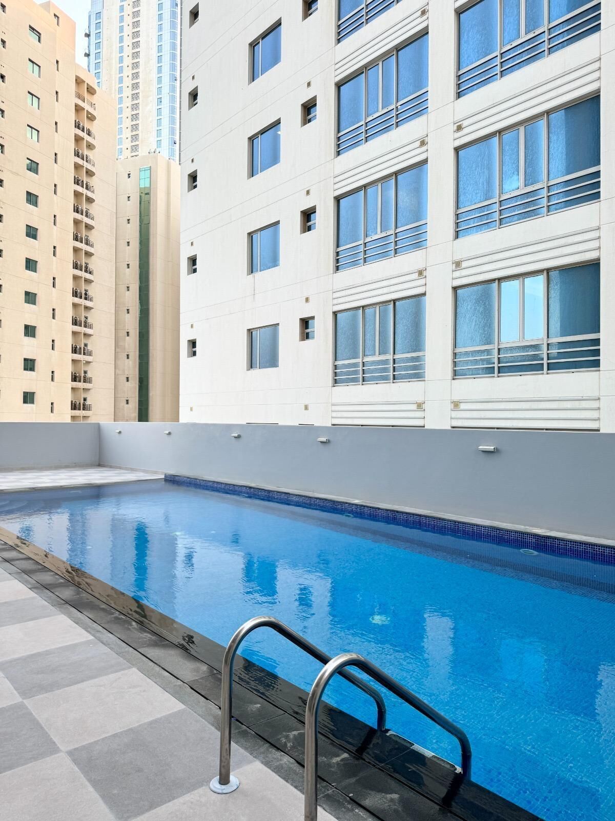 Una piscina con escalera metálica, rodeada por un suelo a cuadros y contigua a un edificio con balcones y ventanas grandes.