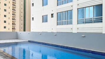 Una piscina con escalera metálica, rodeada por un suelo a cuadros y contigua a un edificio con balcones y ventanas grandes.