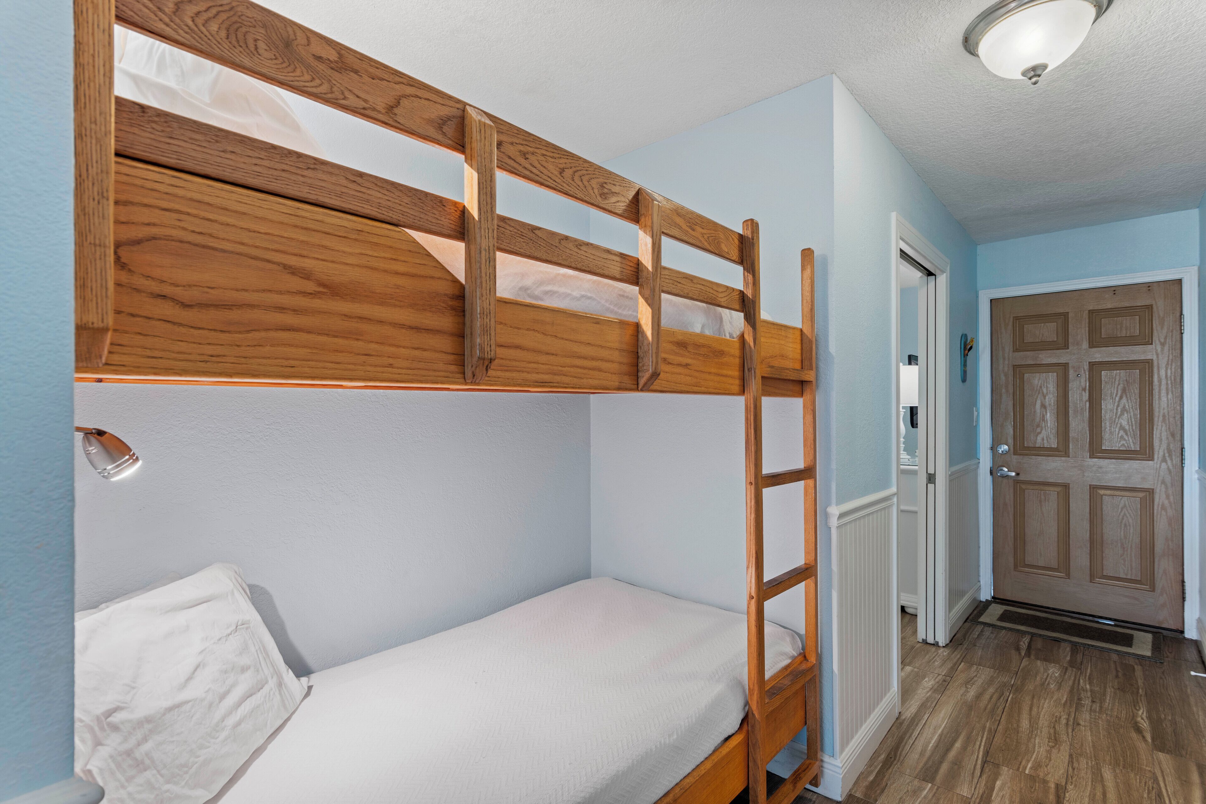 A bunk bed with a wooden ladder, a white mattress, and a white pillow. There is a door with a wooden panel design and a white doormat on the floor.