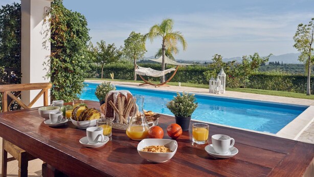 Une table en bois, dressée pour le petit-déjeuner, avec vue sur une piscine et des palmiers.