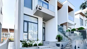 Une maison moderne à deux étages, dotée d’un toit plat, de grandes fenêtres et d’un escalier en béton menant à l’entrée.