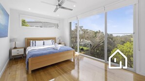 A modern bedroom with a large bed, wooden flooring, a ceiling fan, and a view of the outdoors through large windows. - Banksia Beach Retreat *1 NIGHT FREE* (Blairgowrie)