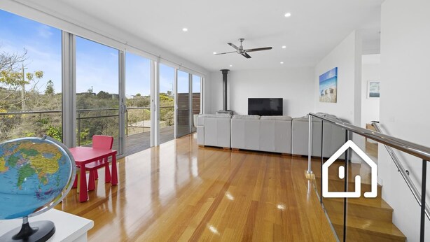 A spacious living area with a large sliding glass door leading to a balcony, a ceiling fan, a flat-screen TV, and a dining area with a red chair. - Banksia Beach Retreat *1 NIGHT FREE* (Blairgowrie)