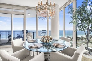 A dining area with a glass table, white chairs, and a chandelier. It features a large window with a view of the ocean and a balcony with chairs.