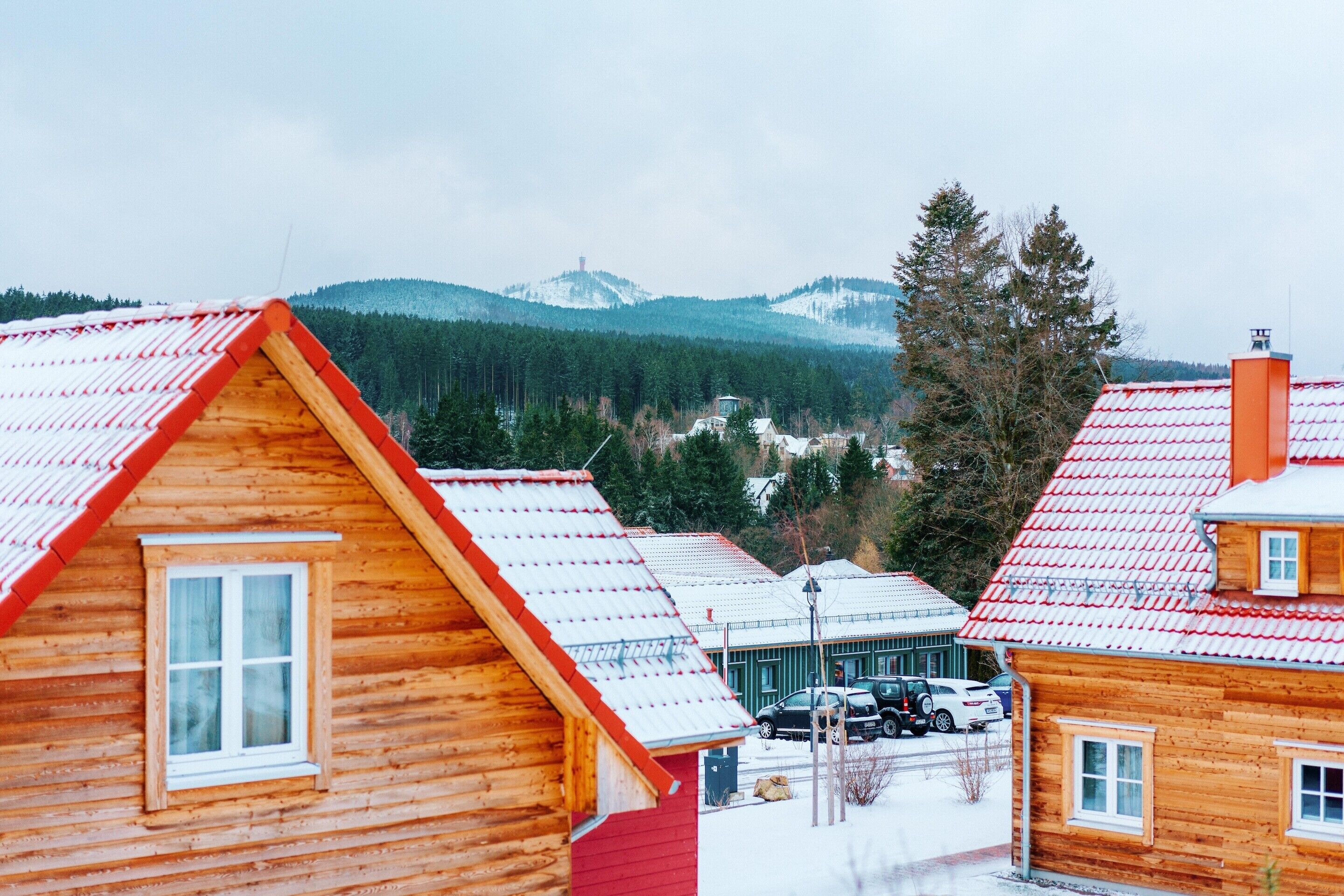 A wooden cabin with a red roof, surrounded by other cabins and a snowy landscape with mountains in the background.