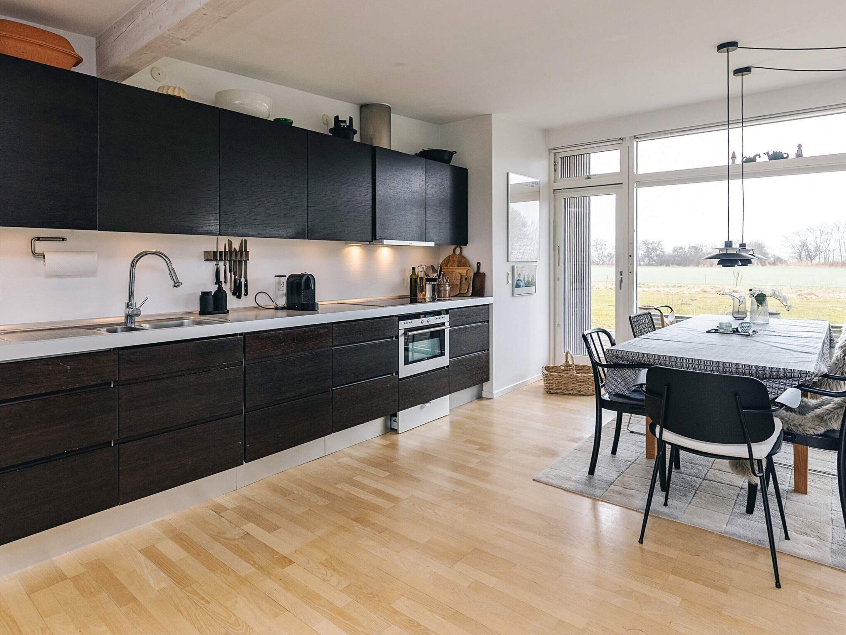 A modern kitchen with dark cabinets, a stainless steel sink, and an oven. There's a dining area with a table and chairs, and a large window offering a view of the outdoors.
