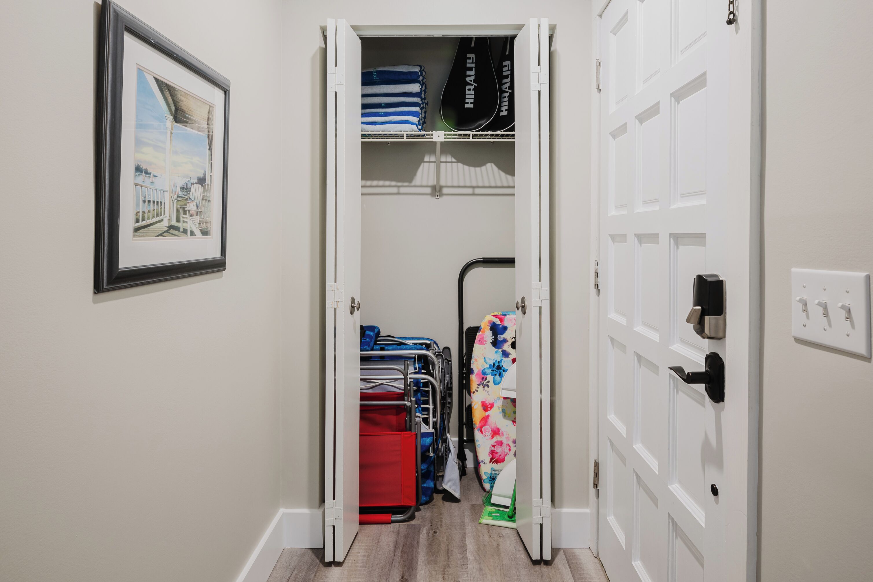 A hallway with a door leading to a room, a framed picture on the wall, and a rack with folded towels and a floral shower curtain.