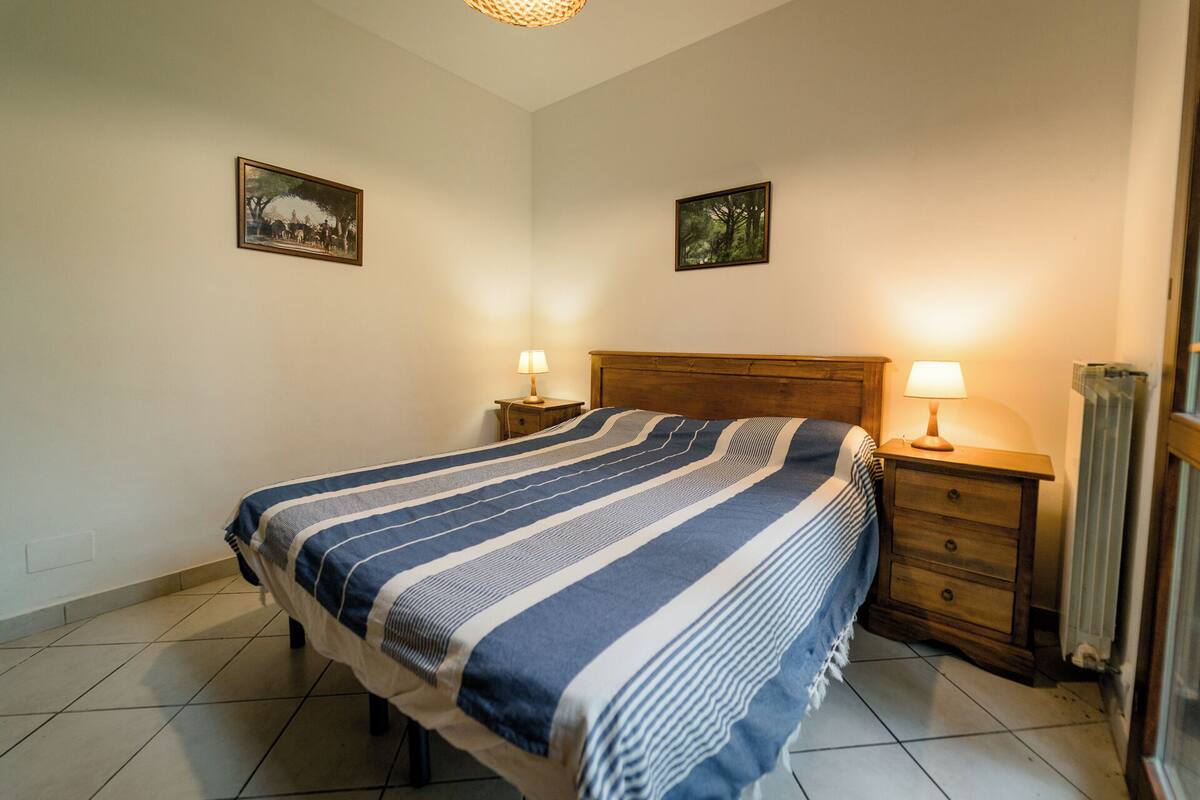 A bedroom with a bed, two bedside tables with lamps, a wooden headboard, and two framed pictures on the wall.