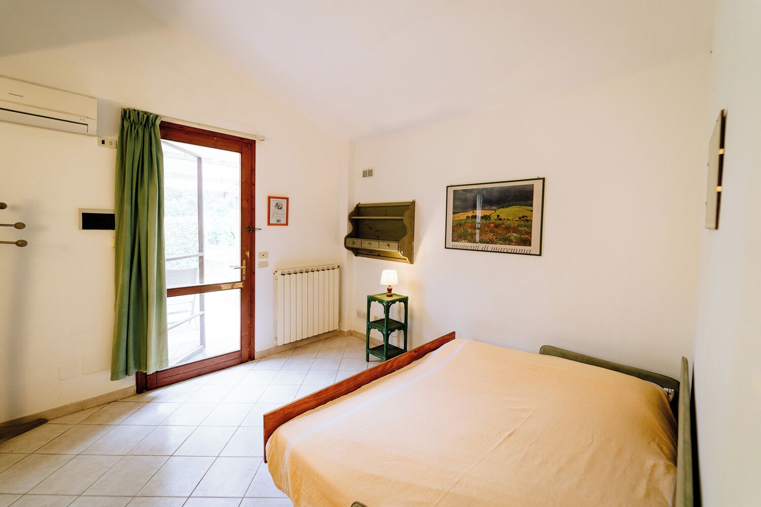 A bedroom with a bed, a radiator, a window with curtains, a wall-mounted shelf, and a framed picture.