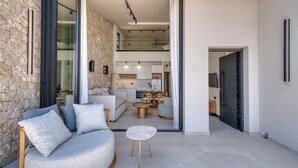 Een moderne zithoek met een stenen muur, een ronde grijze bank met kussens, een klein rond tafeltje en een keuken op de achtergrond.