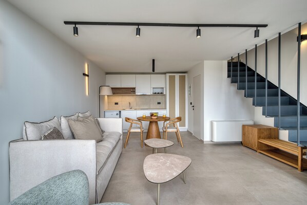 A modern living room with a sofa, a coffee table, and a dining area with chairs. A staircase with a black railing is visible to the right. - Anima Deluxe Suites (Nafplio)