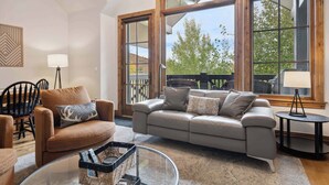 A living room with a sofa, armchair, and coffee table, large windows with a view of trees, and a dining area with chairs and a lamp. - Beaver Creek Landing A408 (Beaver Creek)