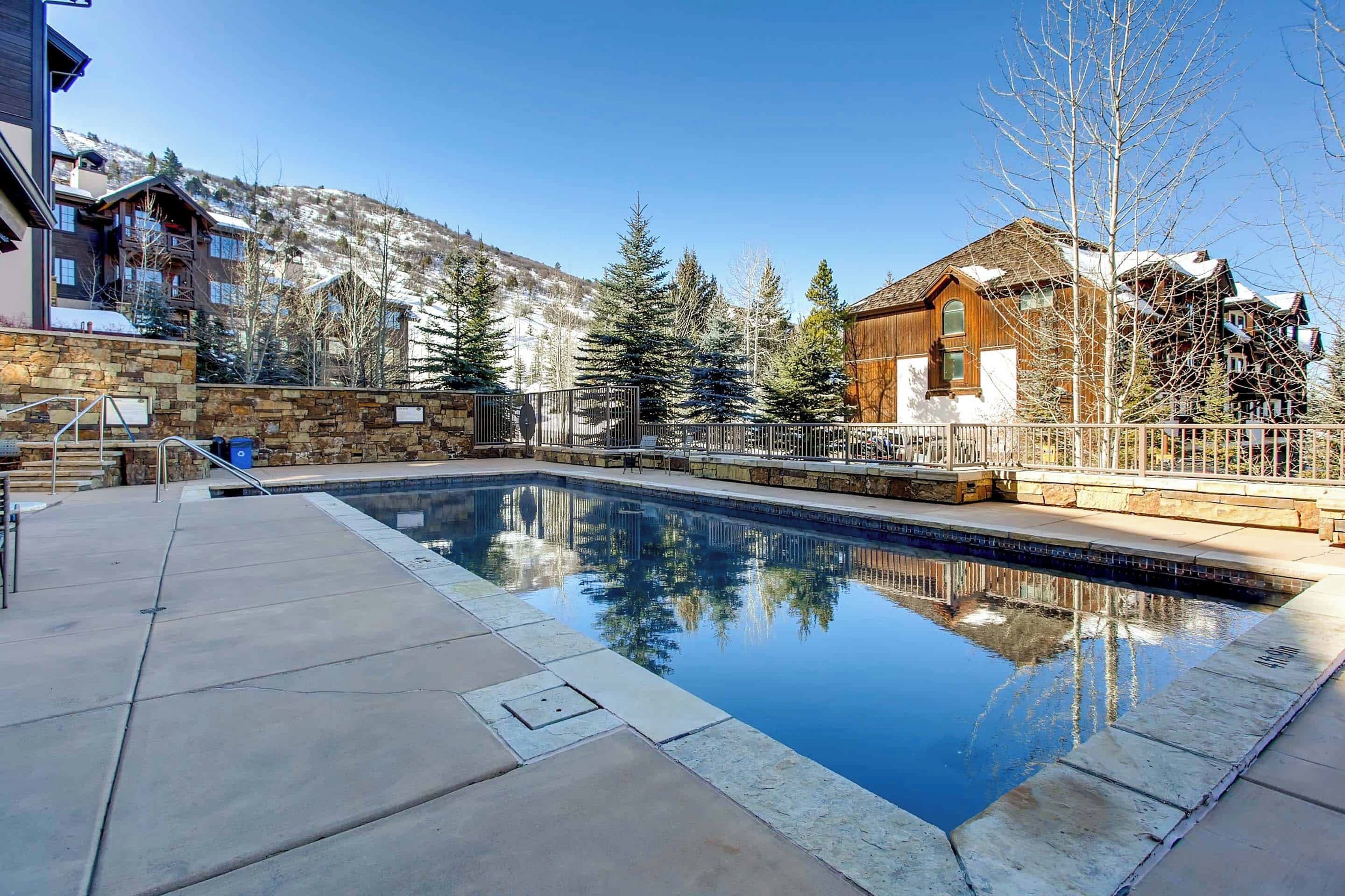 A swimming pool with a stone deck, surrounded by a wooden fence and buildings with a mountainous backdrop.