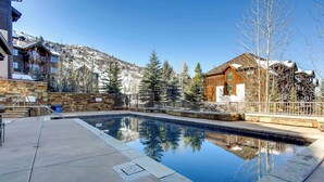 A swimming pool with a stone deck, surrounded by a wooden fence and buildings with a mountainous backdrop. - Beaver Creek Landing A408 (Beaver Creek)
