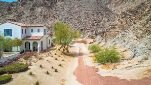 Un edificio blanco con techo de tejas rojas, un sendero de tierra y una montaña rocosa al fondo.