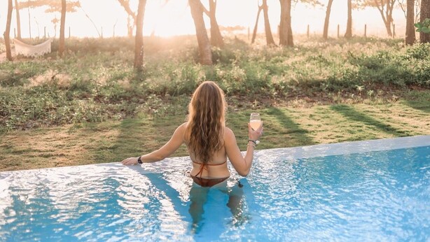 A woman in a bikini at the edge of a pool with a drink, surrounded by trees and a hammock in the background. - Oceanfront Paradise:4-Bedroom Villa with Expansive Pool in Santa Teresa Sea Wing (Puntarenas Province)
